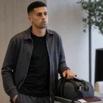 João Cancelo in neutrale reiskleding in een rustige vertrekruimte met een reistas.