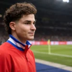 Julián Álvarez in een rode sportjas aan de rand van een voetbalveld.