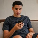 Jamal Musiala kijkt rustig naar zijn smartphone in een neutrale binnenruimte.