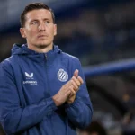 Een Club Brugge-coach in blauwe clubjas applaudisseert langs het veld.