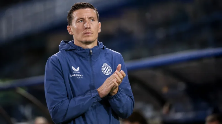 Een Club Brugge-coach in blauwe clubjas applaudisseert langs het veld.