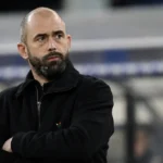 Een niet-spelende voetbalfiguur applaudisseert langs de zijlijn van een stadion.