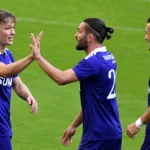 Drie voetballers in paarse wedstrijdkleding tijdens een gezamenlijke opwarming op het veld.