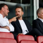 Drie clubofficials in formele kleding in een voetbaltribune.