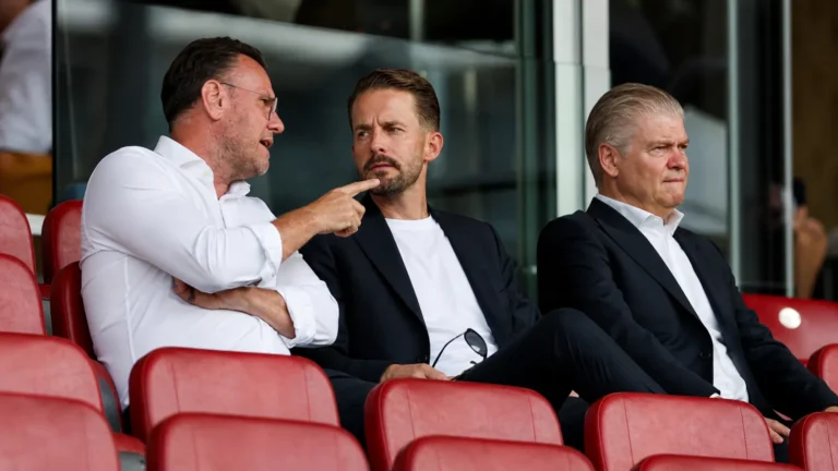 Drie clubofficials in formele kleding in een voetbaltribune.