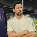 Een coach in een licht trainingsshirt kijkt tijdens de opwarming vanaf de technische zone over het veld, met een Anderlecht-embleem op de dug-out.