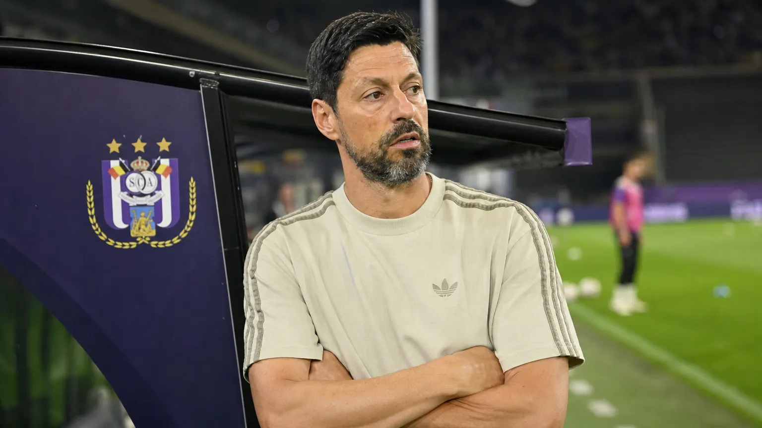 Een coach in een licht trainingsshirt kijkt tijdens de opwarming vanaf de technische zone over het veld, met een Anderlecht-embleem op de dug-out.
