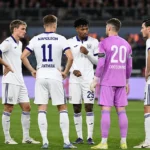 Een groep Anderlecht-spelers in witte wedstrijdtenues overlegt vlak voor een wedstrijd op het veld.