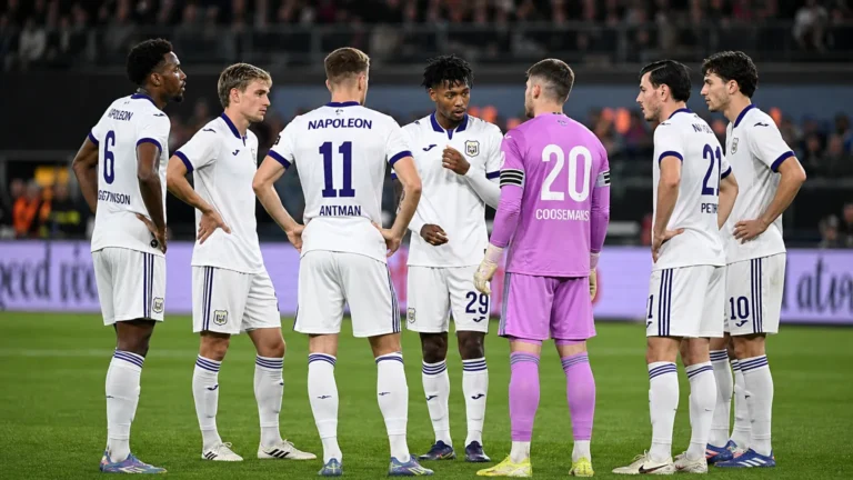 Een groep Anderlecht-spelers in witte wedstrijdtenues overlegt vlak voor een wedstrijd op het veld.