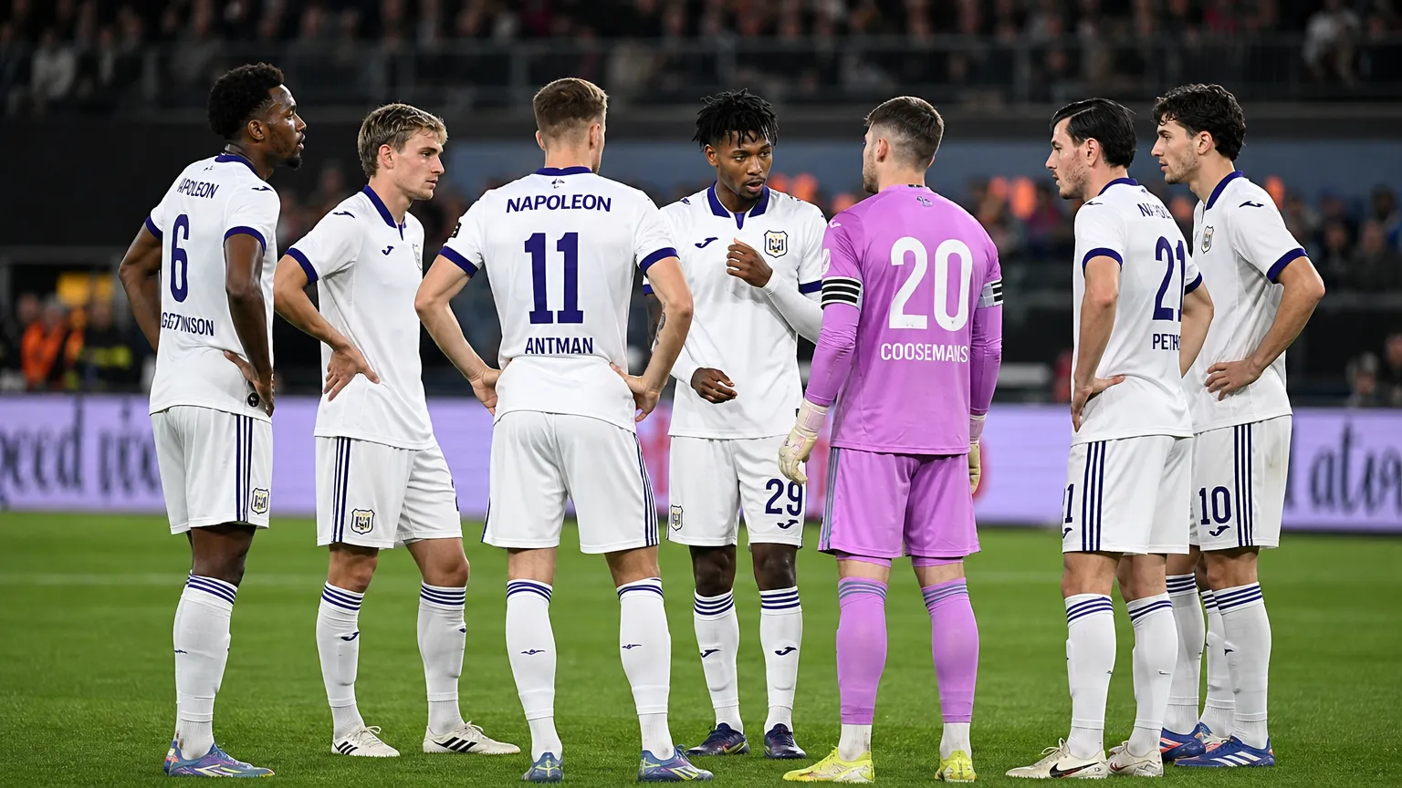 Een groep Anderlecht-spelers in witte wedstrijdtenues overlegt vlak voor een wedstrijd op het veld.