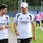 Twee leden van de technische staf van Olympique Lyon overleggen naast een trainingsveld.