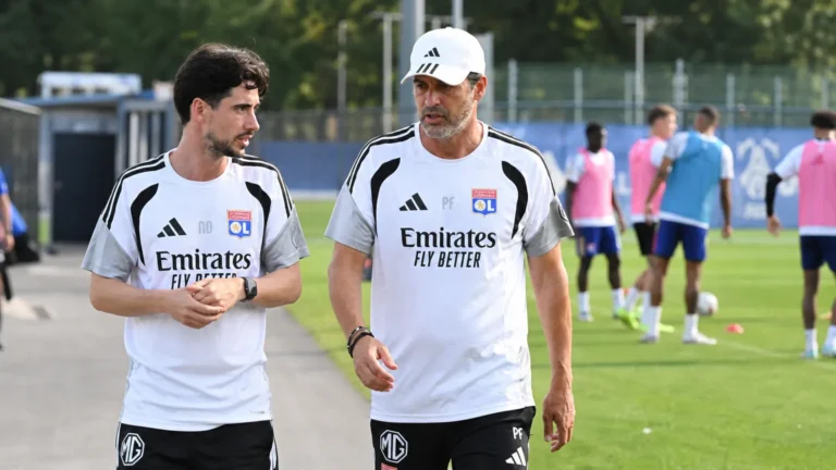 Twee leden van de technische staf van Olympique Lyon overleggen naast een trainingsveld.