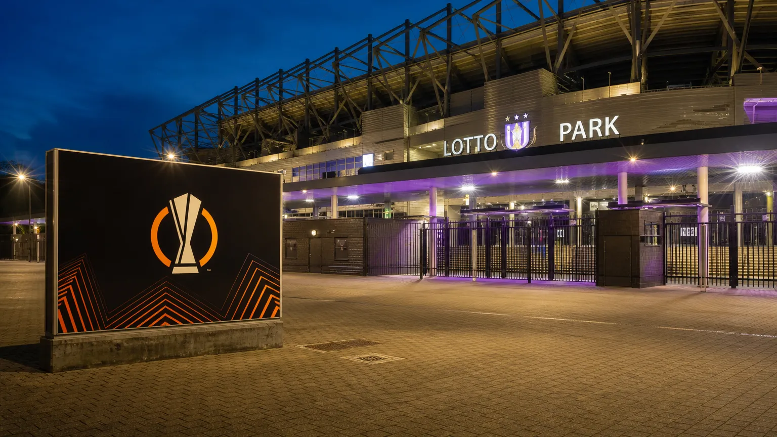 Verlichte ingang van het Lotto Park met een fysiek Europa League-wedstrijdbord.