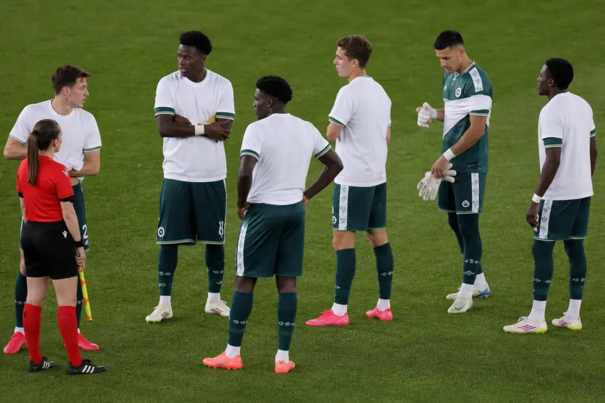 Een groep spelers en een official op het veld voor de aftrap, met witte steunshirts over wedstrijdkleding.