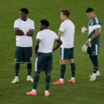 Een groep spelers en een official op het veld voor de aftrap, met witte steunshirts over wedstrijdkleding.