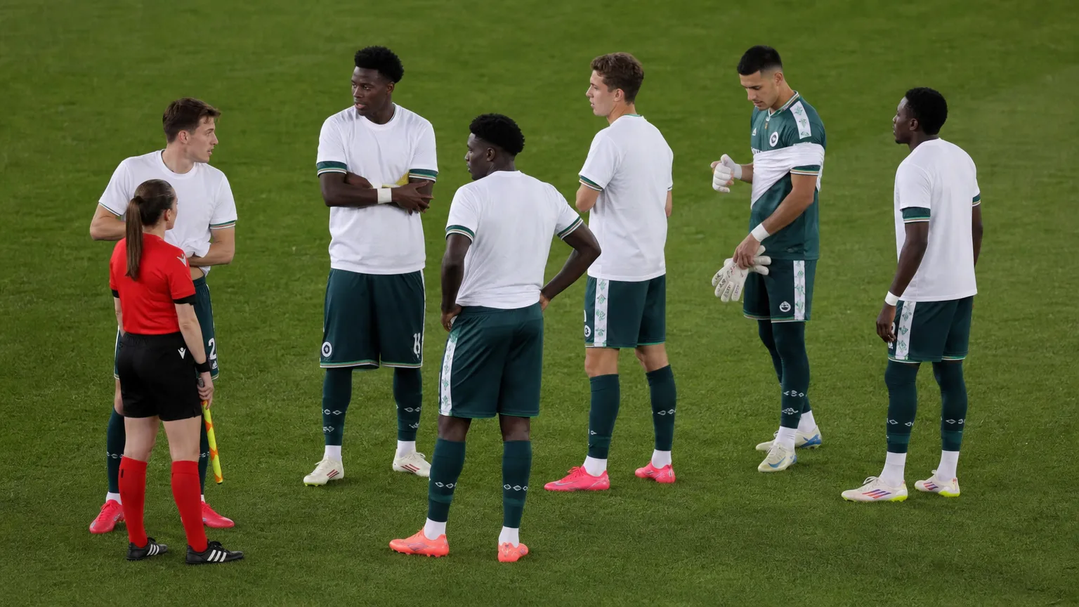 Een groep spelers en een official op het veld voor de aftrap, met witte steunshirts over wedstrijdkleding.
