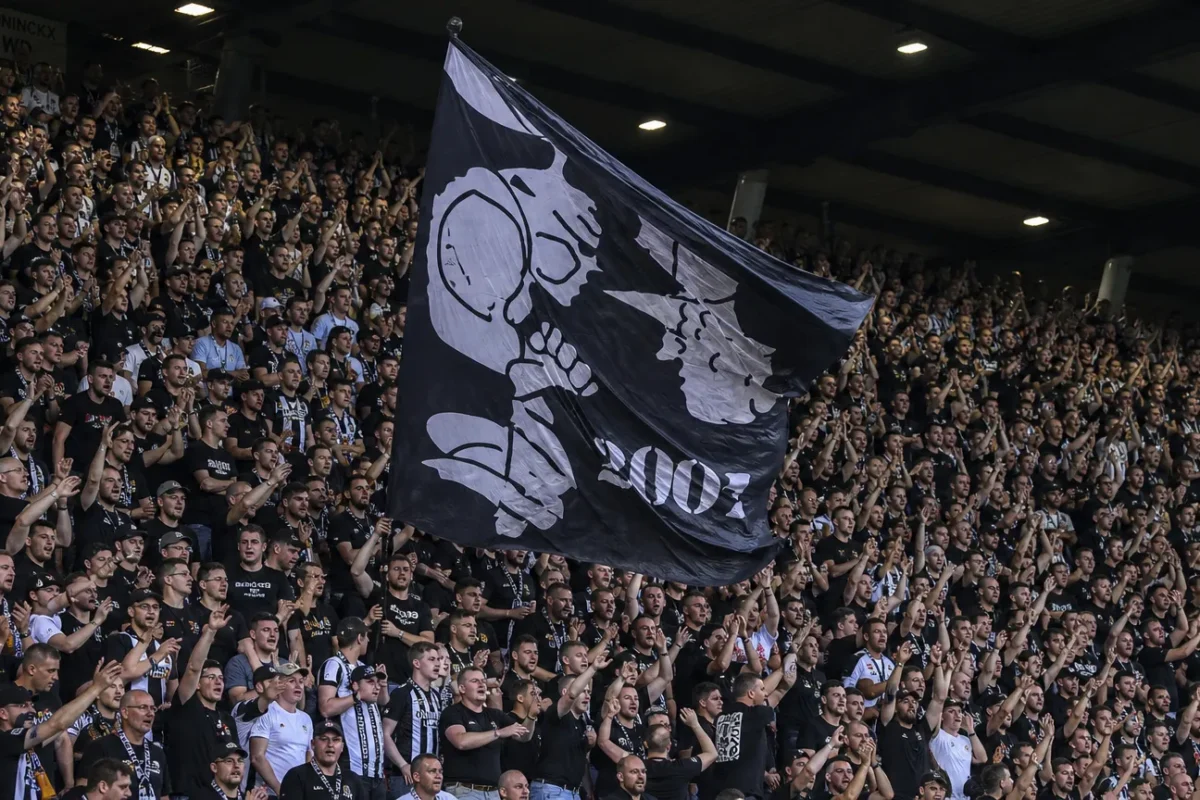 Een volle tribune met supporters in zwart-witte kleding die een grote vlag omhooghouden.