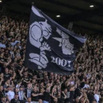 Een volle tribune met supporters in zwart-witte kleding die een grote vlag omhooghouden.