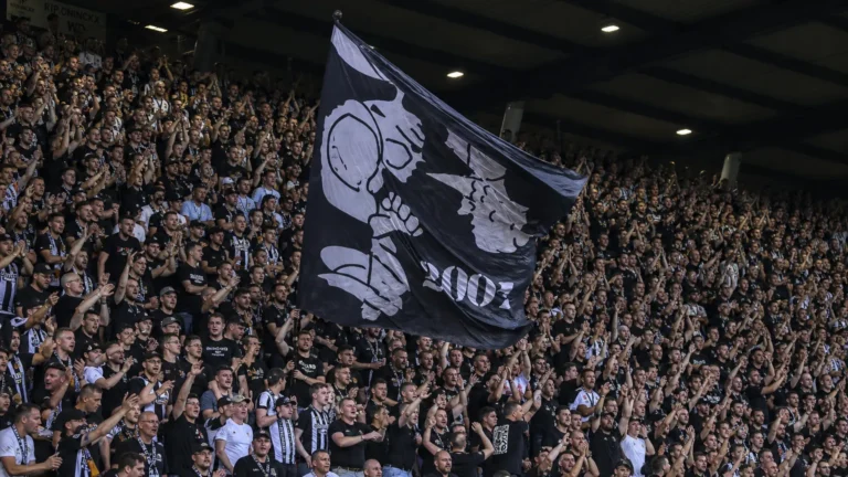 Een volle tribune met supporters in zwart-witte kleding die een grote vlag omhooghouden.