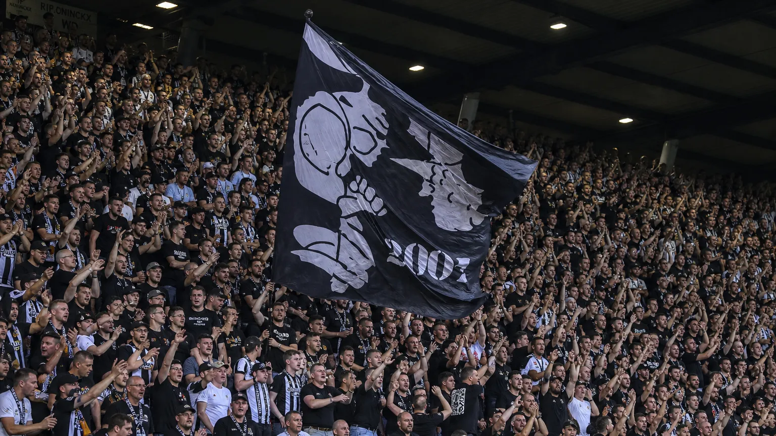 Een volle tribune met supporters in zwart-witte kleding die een grote vlag omhooghouden.