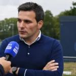 Een niet-spelende clubvertegenwoordiger wordt geïnterviewd naast een voetbalveld met een Union-embleem op een vast bord.