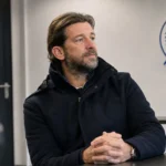 Rik De Mil in een donkere jas in een clubmedia-ruimte met een discreet KAA Gent-wanddetail.
