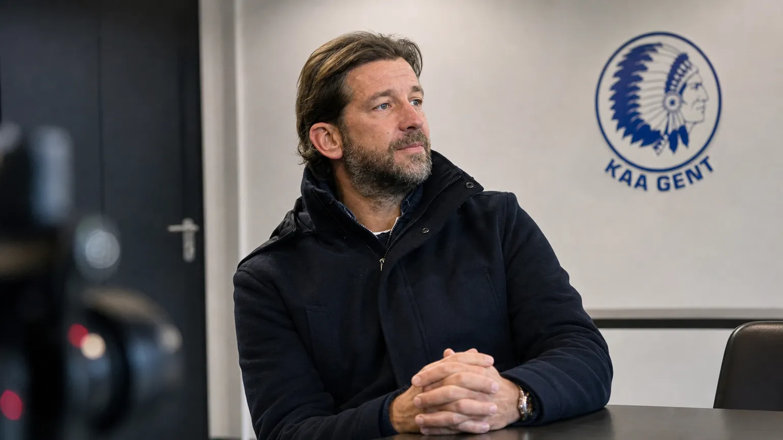 Rik De Mil in een donkere jas in een clubmedia-ruimte met een discreet KAA Gent-wanddetail.