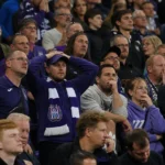 Een collectieve groep Anderlecht-supporters in paars-witte kledij op de tribune met een sjaal van de club.