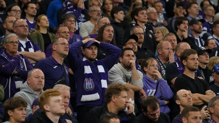 Een collectieve groep Anderlecht-supporters in paars-witte kledij op de tribune met een sjaal van de club.