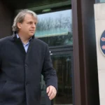 Todd Boehly in donkere jas bij de ingang van een Chelsea-clubgebouw.