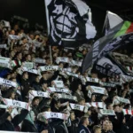 Een groep OHL-supporters zwaait met sjaals en vlaggen in een volle tribune.