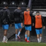 Voetballers in trainingskleding lopen door hevige regen richting een ondergelopen spelerstunnel.