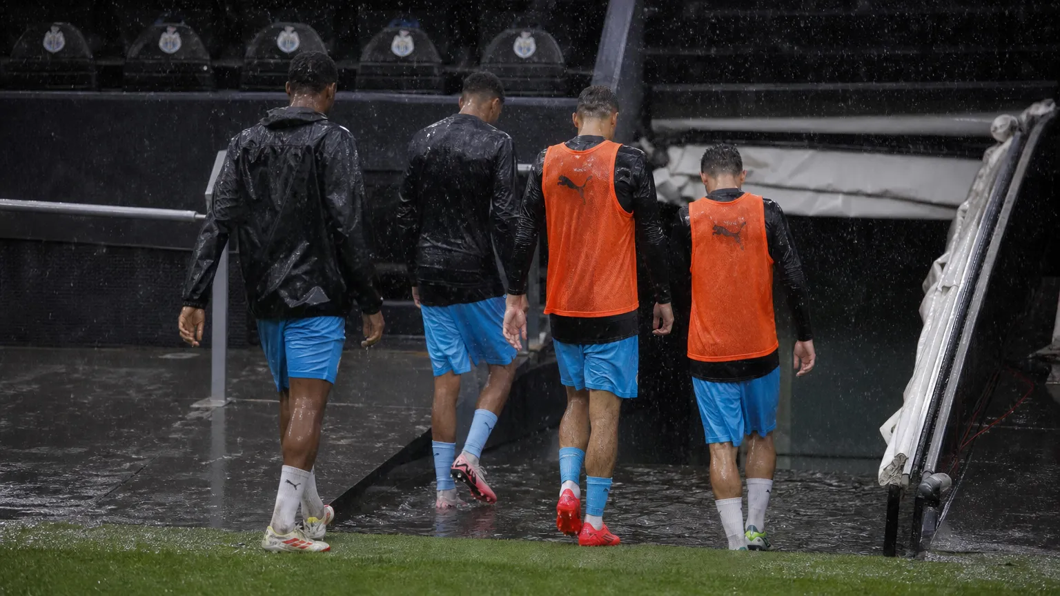 Voetballers in trainingskleding lopen door hevige regen richting een ondergelopen spelerstunnel.