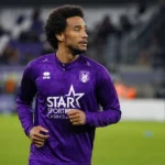 Ryan Sanusi warmt op in een paars voetbaltenue voor een verlichte tribune.