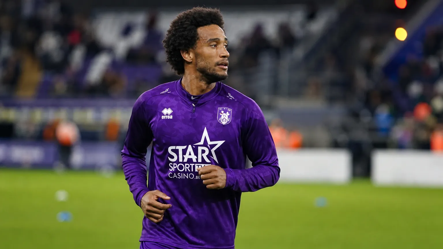 Ryan Sanusi warmt op in een paars voetbaltenue voor een verlichte tribune.
