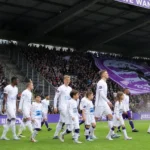 Spelers en kindmascottes lopen het veld op voor een volle overdekte tribune met een groot paars spandoek.