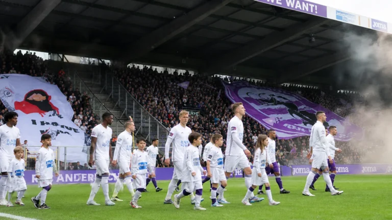 Spelers en kindmascottes lopen het veld op voor een volle overdekte tribune met een groot paars spandoek.