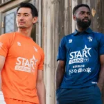 Twee voetballers tonen een oranje en donkerblauw derde shirt tegen een industriële achtergrond.