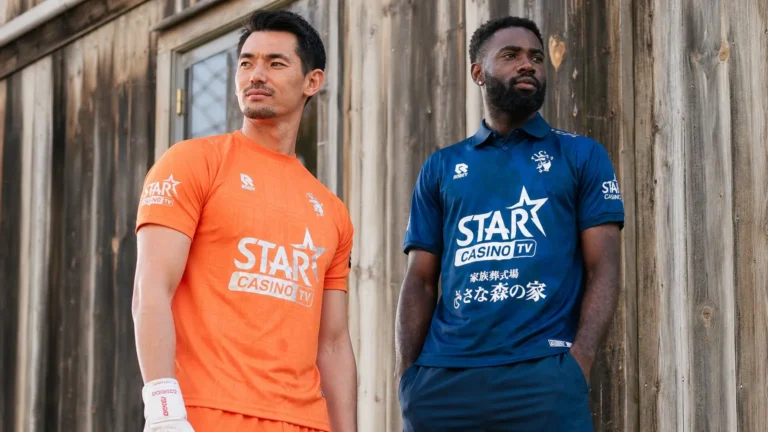 Twee voetballers tonen een oranje en donkerblauw derde shirt tegen een industriële achtergrond.