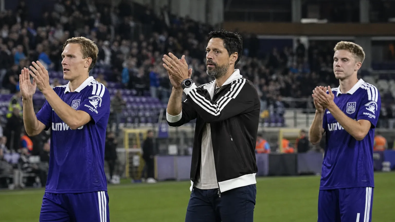 Een niet-spelende clubvertegenwoordiger en spelers in paarse tenues applaudisseren langs het veld.