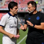 Een Club Brugge-coach overlegt naast het veld met een speler in een wit shirt.