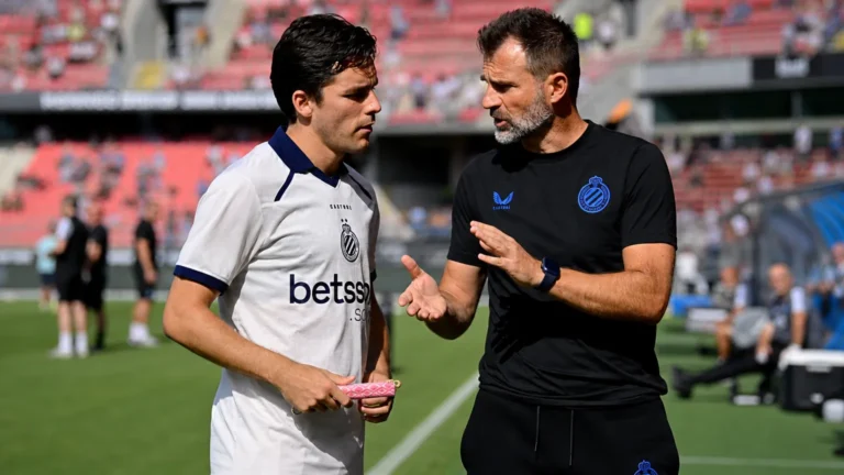 Een Club Brugge-coach overlegt naast het veld met een speler in een wit shirt.