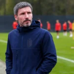 Mark van Bommel in een donkere coachjas langs de zijlijn, met spelers in rode shirts op de achtergrond.