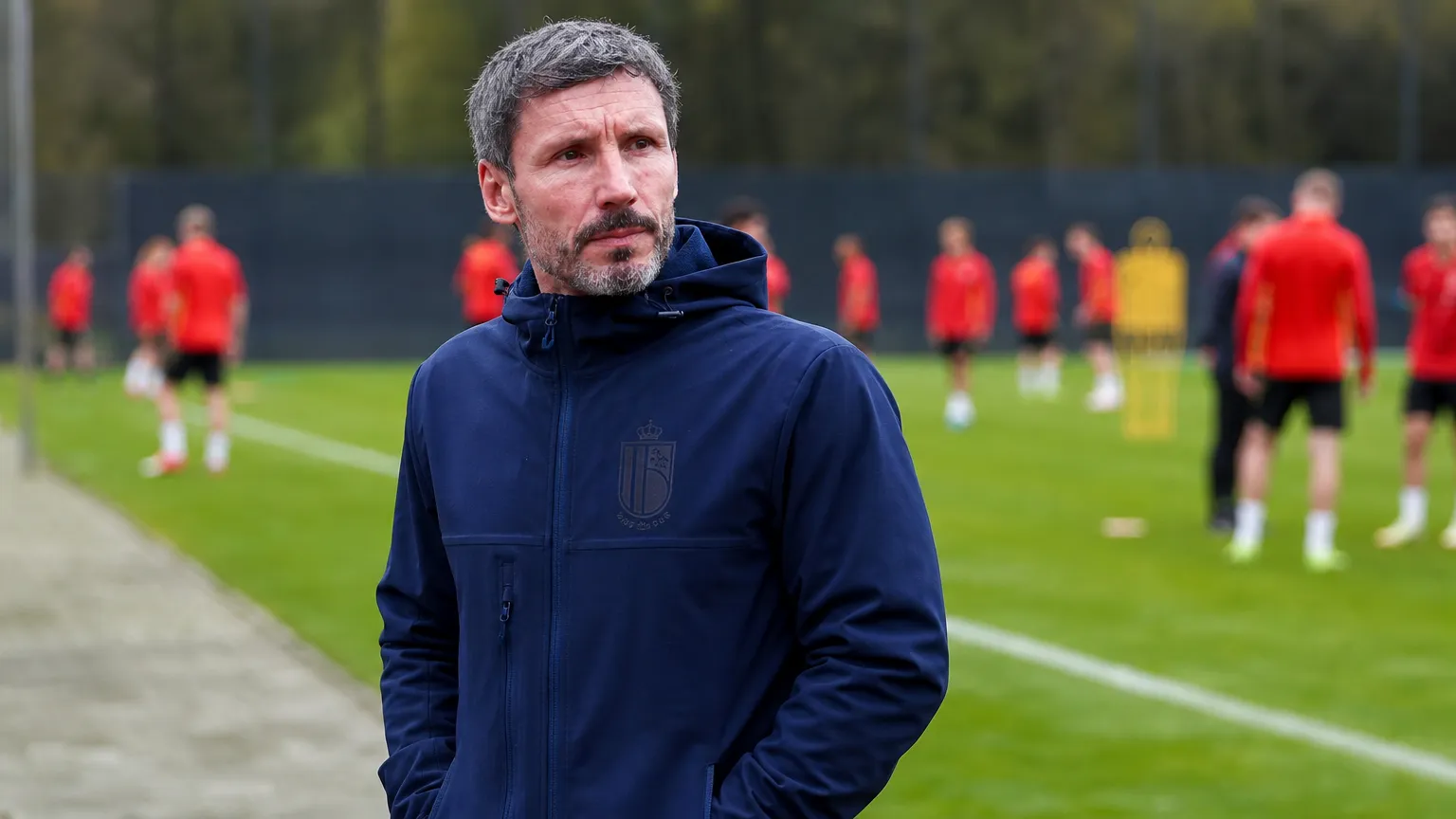 Mark van Bommel in een donkere coachjas langs de zijlijn, met spelers in rode shirts op de achtergrond.