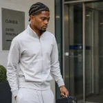 Leon Bailey in neutrale sportkleding bij de ingang van een medische kliniek.