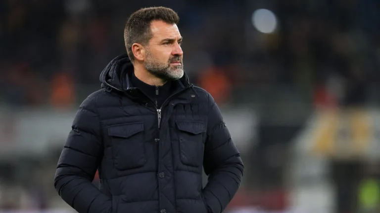 Een coach in donkere jas kijkt aandachtig vanaf de zijlijn van een voetbalveld.