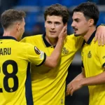 Drie voetballers in gele wedstrijdshirts vieren samen op het veld.