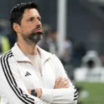 Een coach in een witte sporttop observeert een wedstrijd vanuit de technische zone.