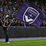 Een Bordeaux-supporter zwaait langs het veld met een grote paarse clubvlag, met een volle tribune op de achtergrond.
