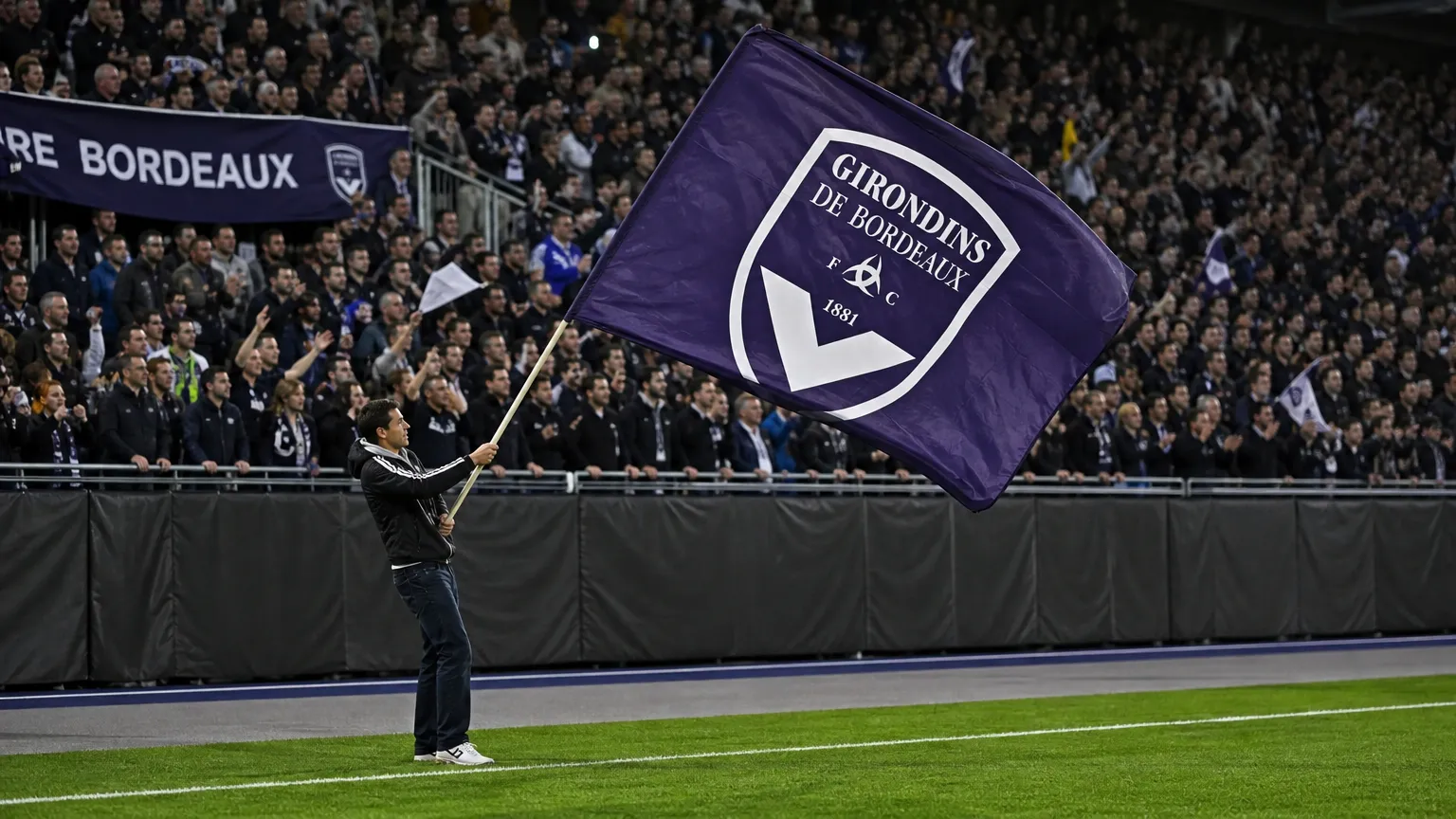 Een Bordeaux-supporter zwaait langs het veld met een grote paarse clubvlag, met een volle tribune op de achtergrond.